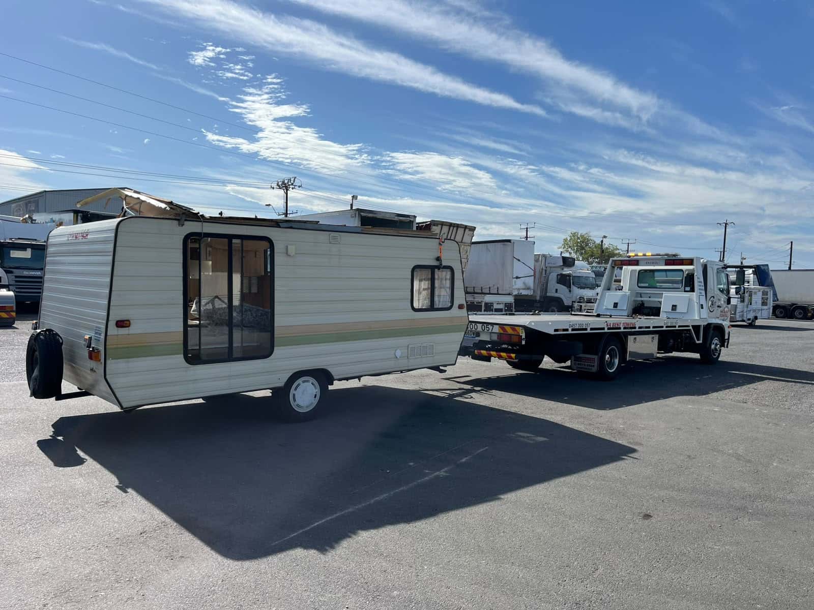 Older cream single-axle caravan with front roof damage on a sealed yard, the SA Best Towing tilt tray parked beyond it