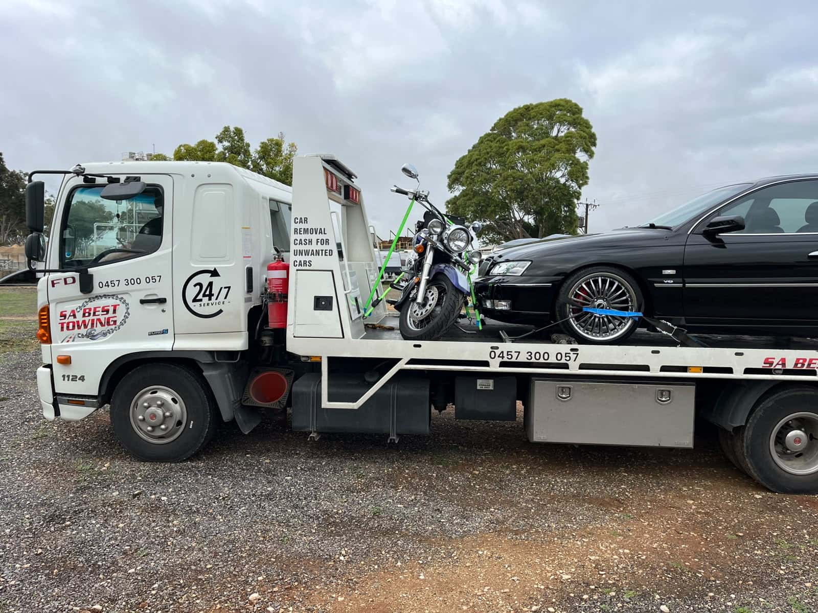Cruiser motorcycle strapped upright behind the headboard and a black Holden sedan on the same tray, gravel yard