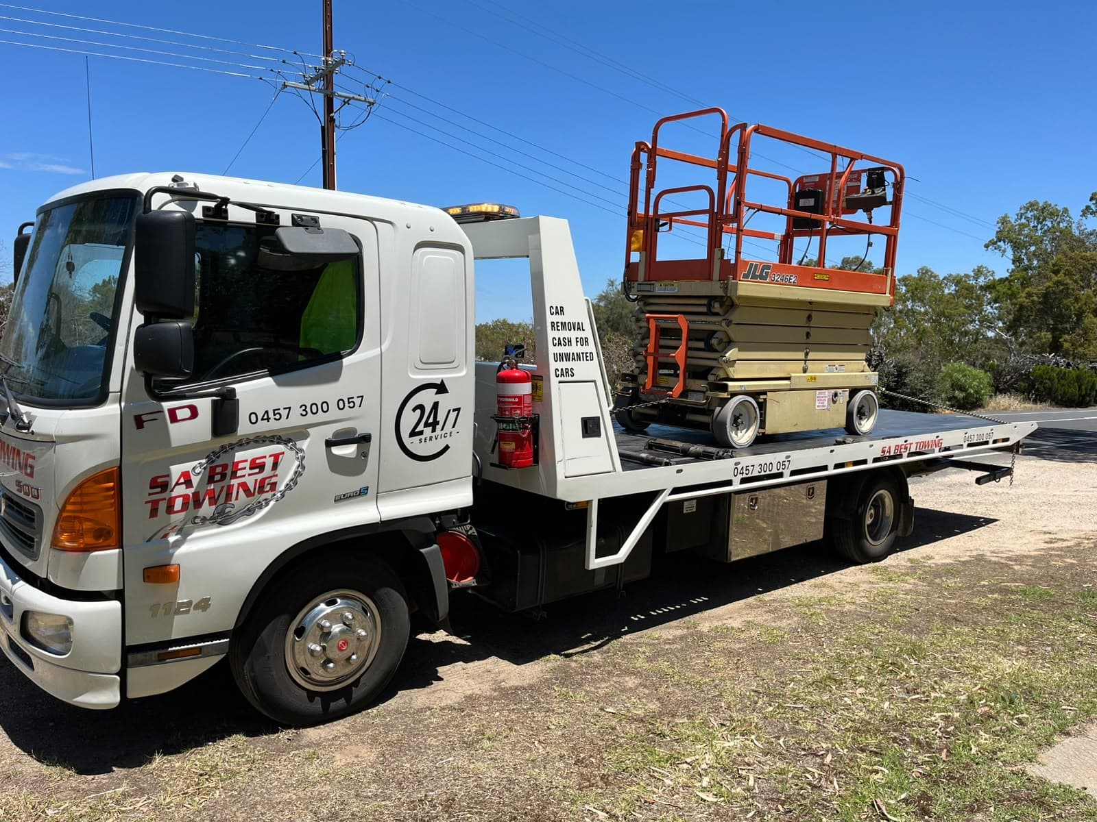 Orange JLG 3246E2 scissor lift with its platform lowered, chained on the SA Best Towing Hino tilt tray beside a rural road