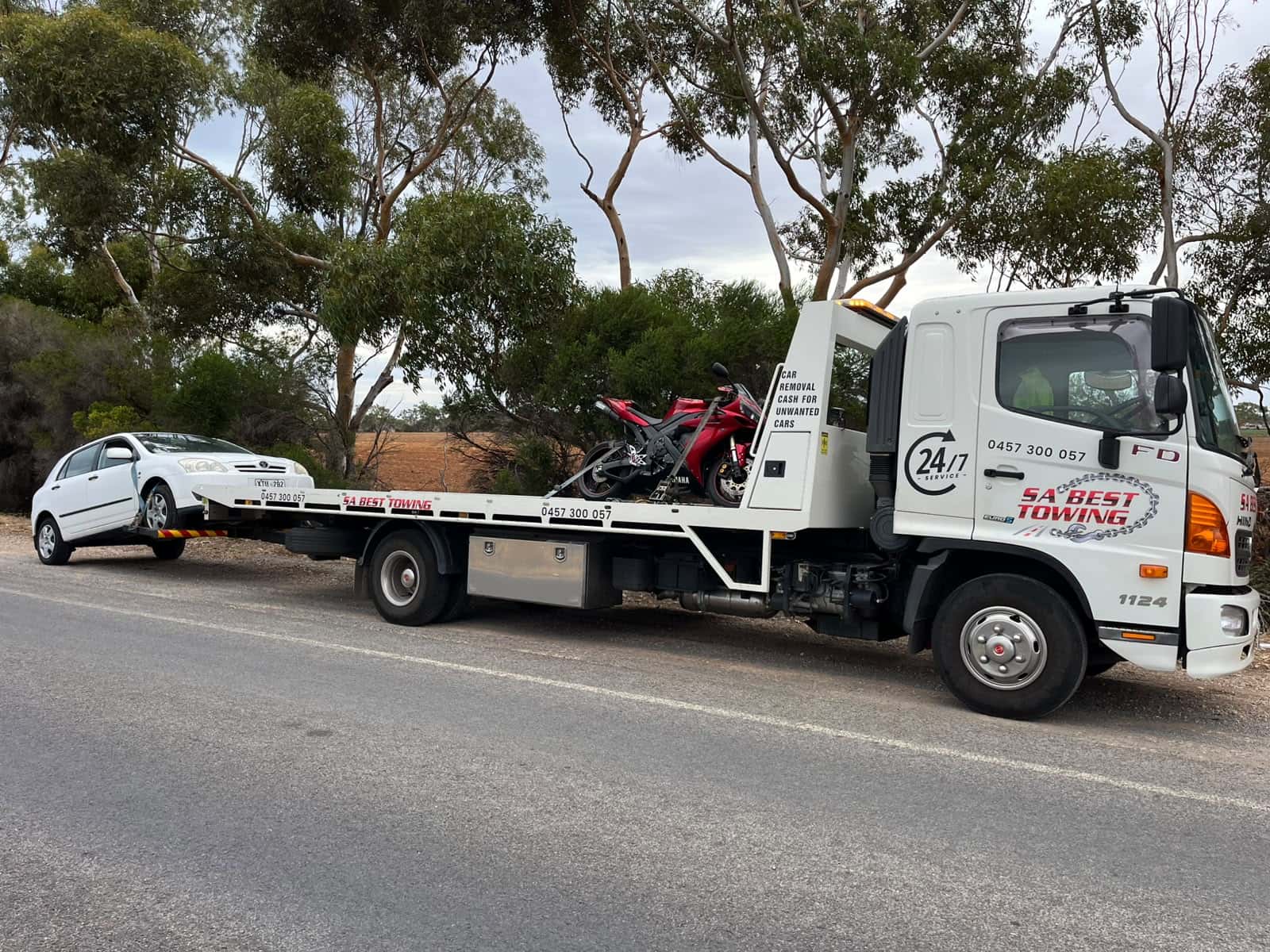 Red and black sports motorcycle strapped on the tray, a white hatchback on the rear lift, gum trees along the road