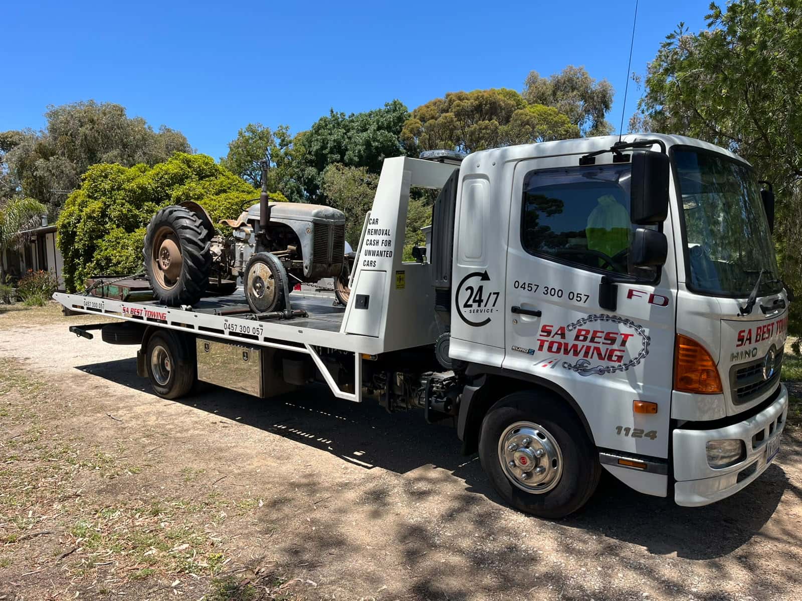 Grey vintage tractor on the SA Best Towing tilt tray seen past the front of the Hino cab, garden and gum trees behind