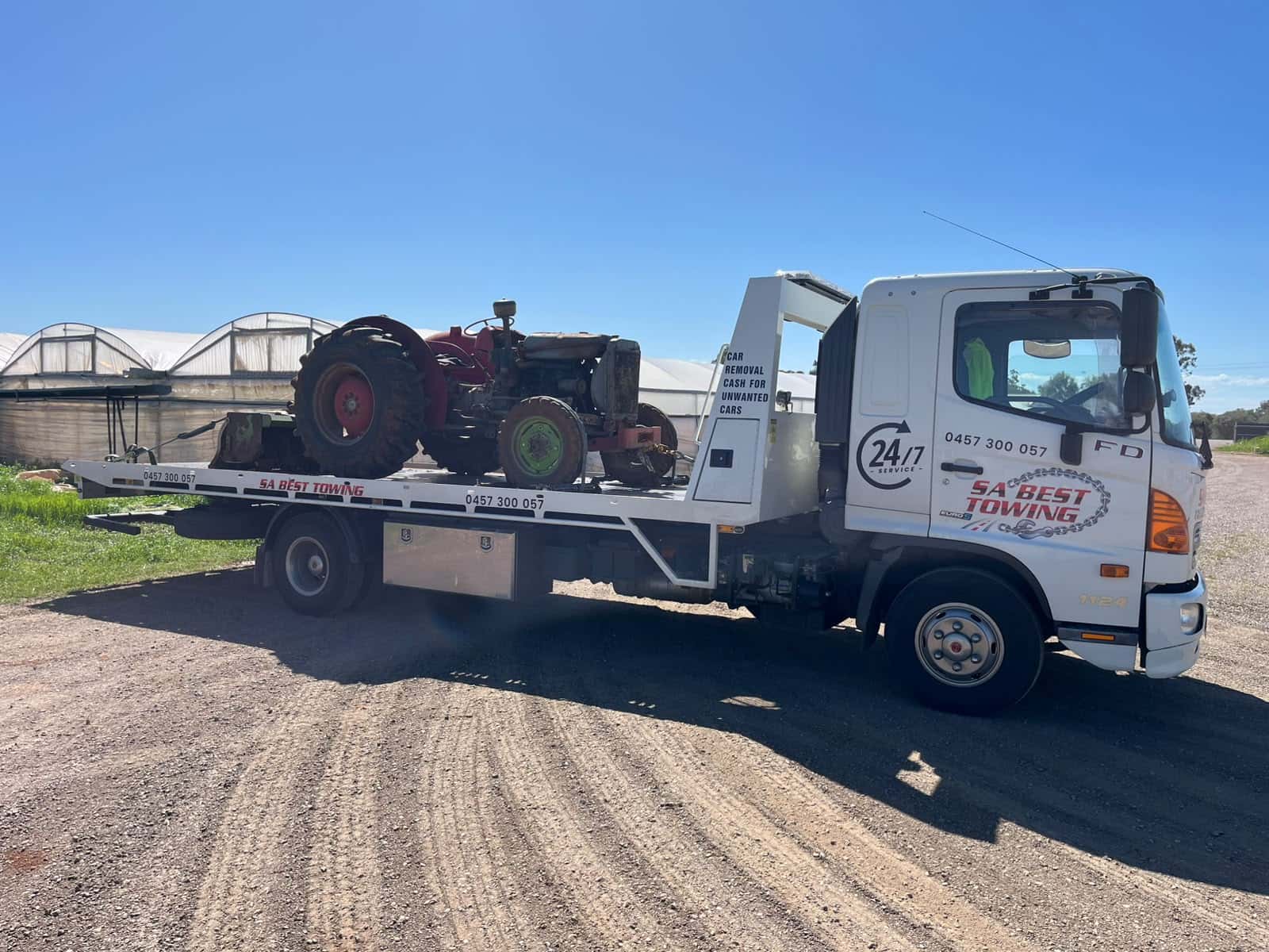 Red vintage tractor with green front wheels chained on the SA Best Towing tilt tray, a green implement strapped behind it, greenhouses in the background